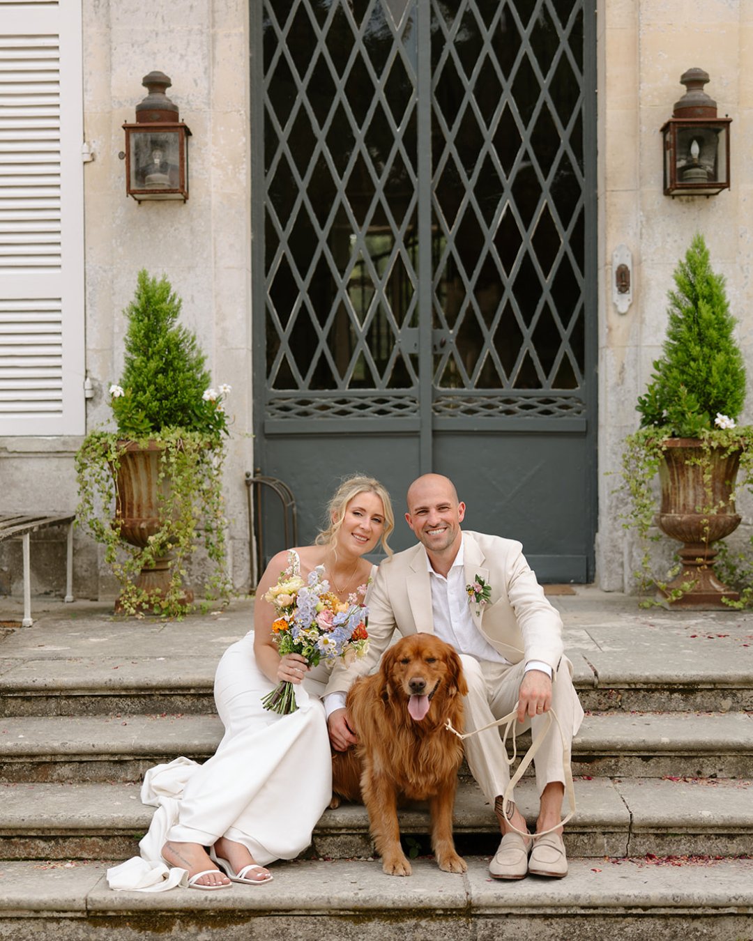 A bride and groom sit on stone steps with a golden retriever between them, smiling in front of a large, ornate door.