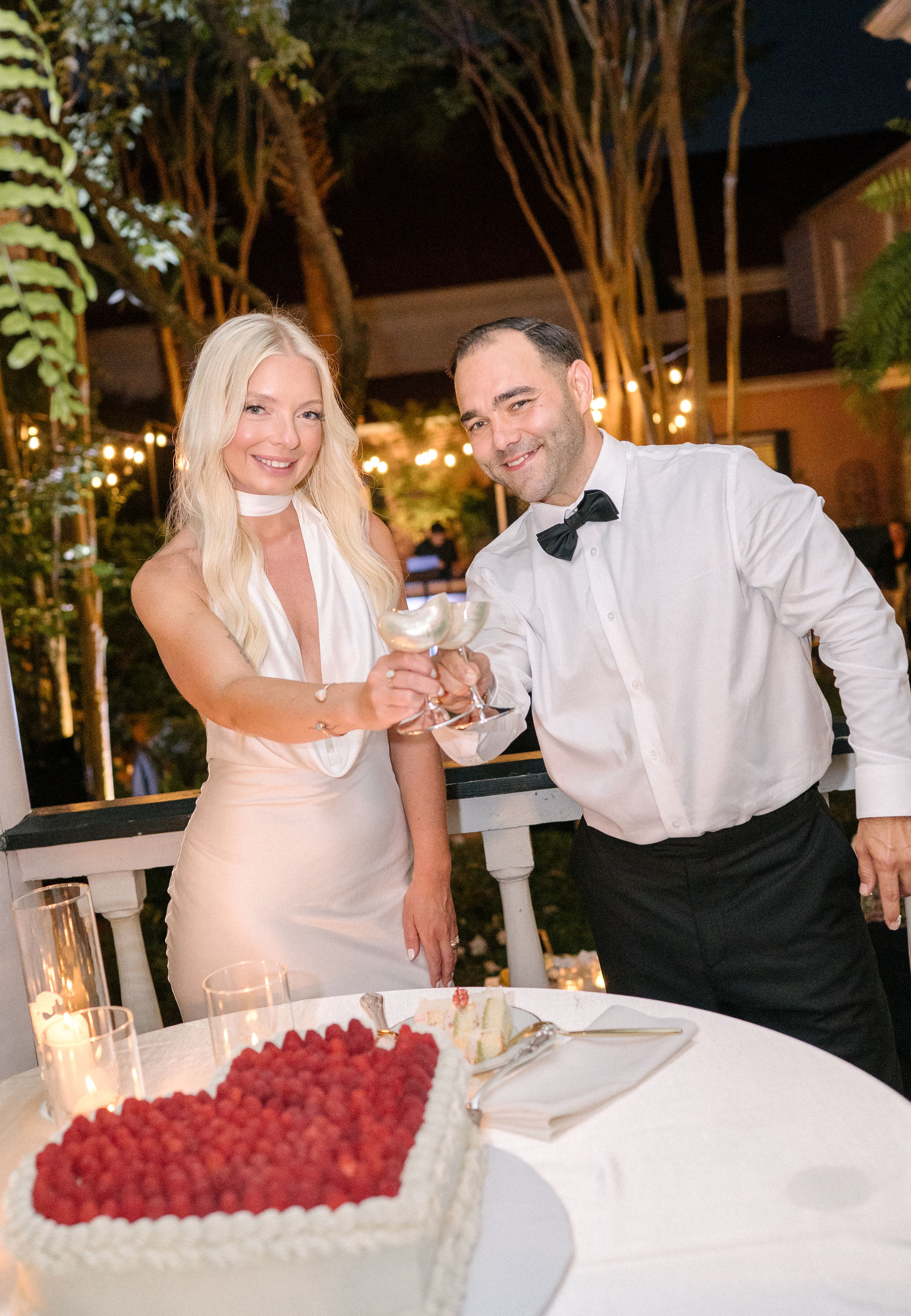 A woman in a white dress and a man in a white shirt with a black bow tie clink glasses beside a heart-shaped cake on a table outdoors at night.