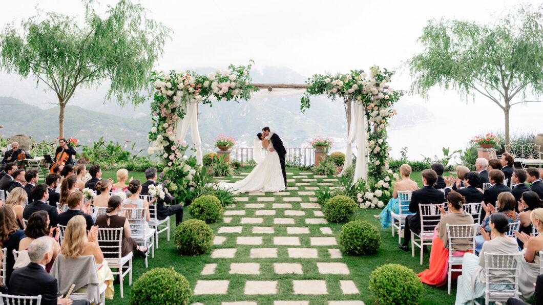 Bride and groom kiss under a flower-adorned arch at an outdoor wedding ceremony, with guests seated on either side of a stone path surrounded by greenery.