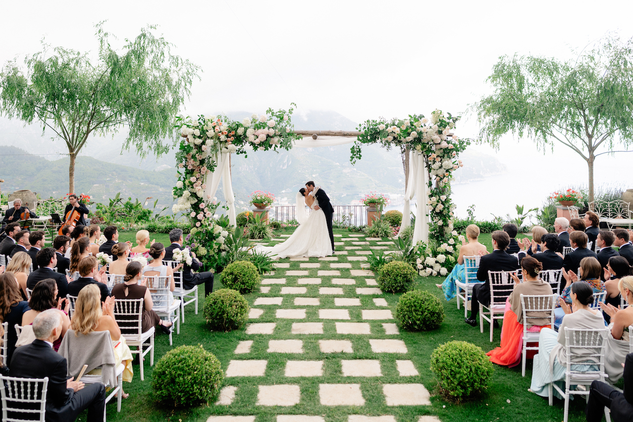 Bride and groom kiss under a flower-adorned arch at an outdoor wedding ceremony, with guests seated on either side of a stone path surrounded by greenery.