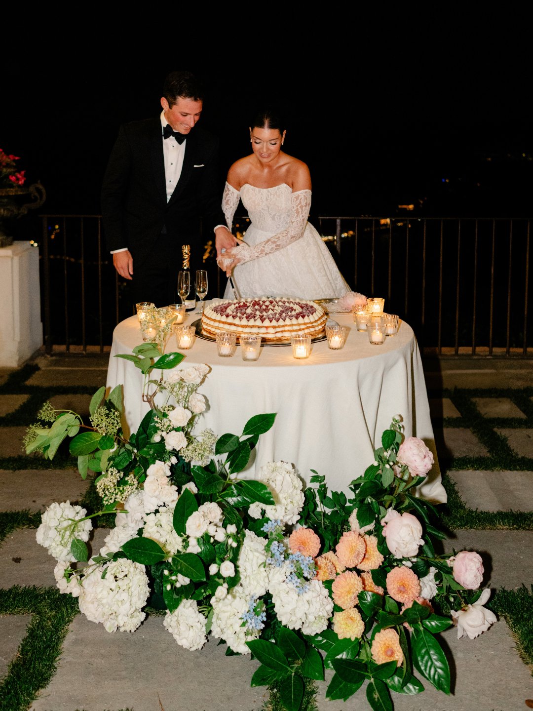 A couple in formal wedding attire cut a cake together at an outdoor table decorated with candles and a large floral arrangement.