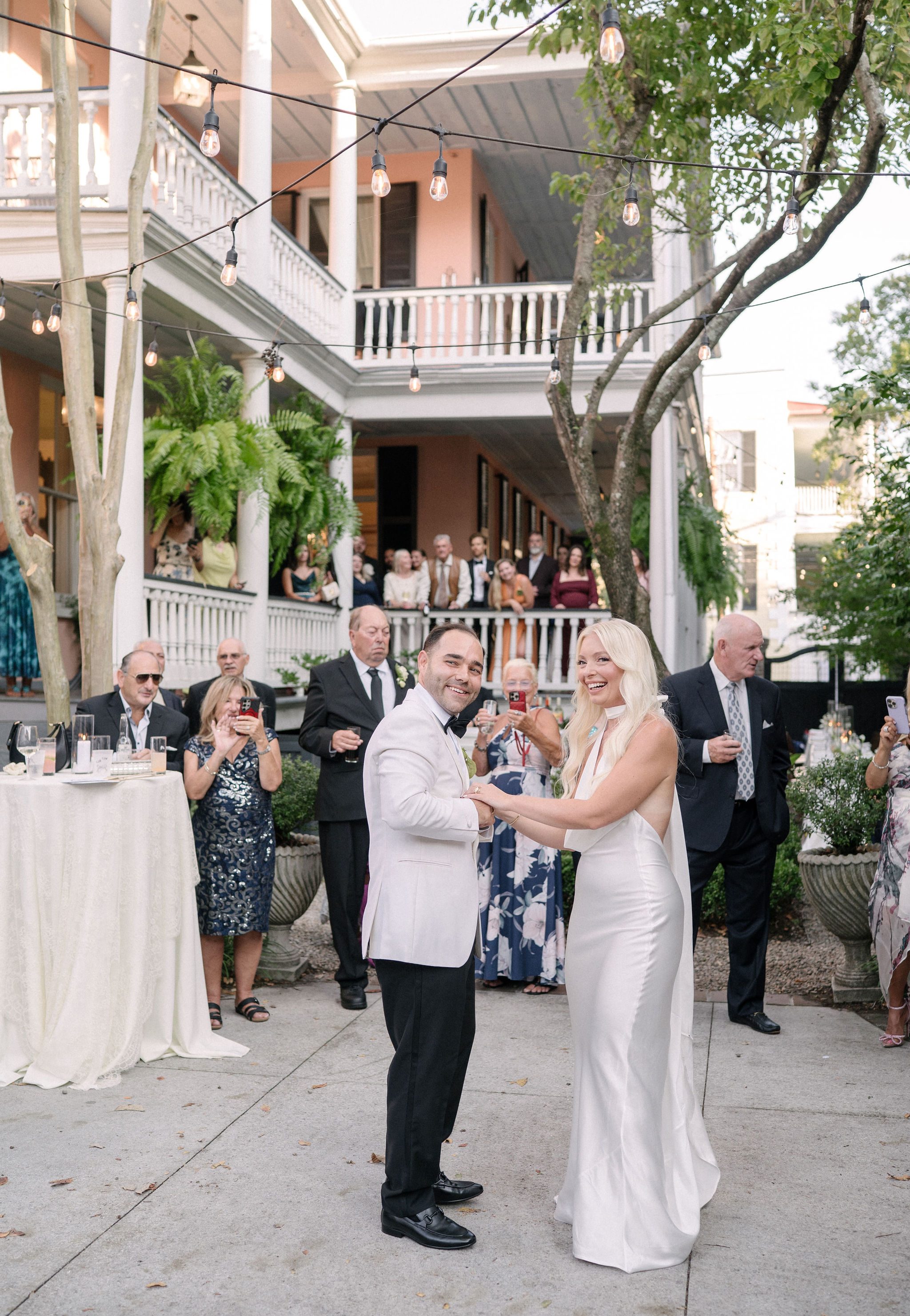 A bride and groom dance outside at their wedding reception, surrounded by guests and string lights in front of a two-story house.