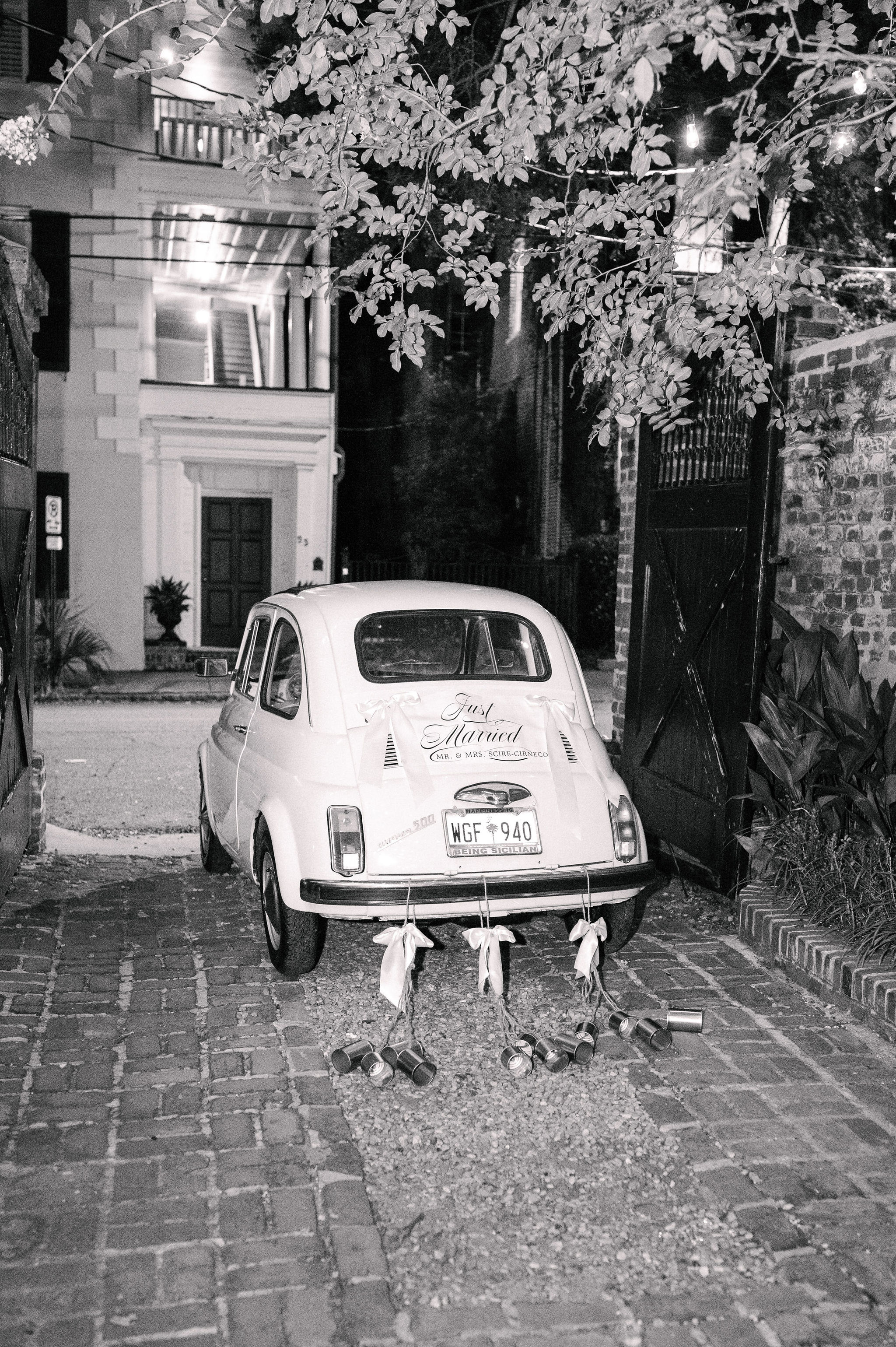 A vintage car with a "Just Married" sign and cans tied to the bumper is parked in a cobblestone driveway at night.