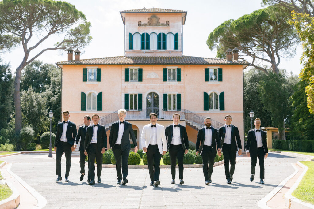 A group of men in formal suits, one in a white jacket, walk in front of a large, elegant villa with green shutters and arched windows.