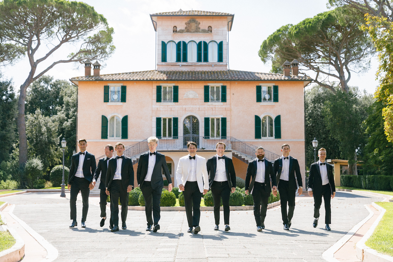A group of men in formal suits, one in a white jacket, walk in front of a large, elegant villa with green shutters and arched windows.