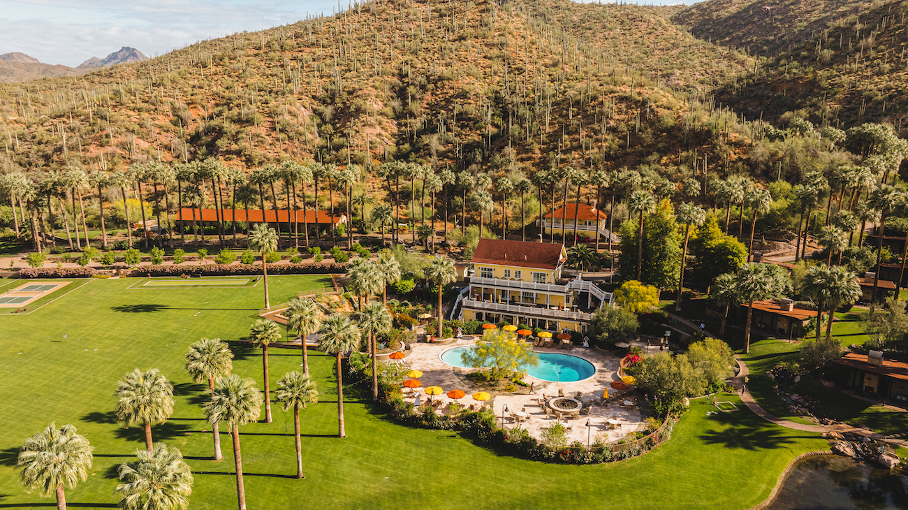 Aerial view of a resort with a round outdoor swimming pool, surrounded by palm trees and lush green lawns, set against a backdrop of desert hills.