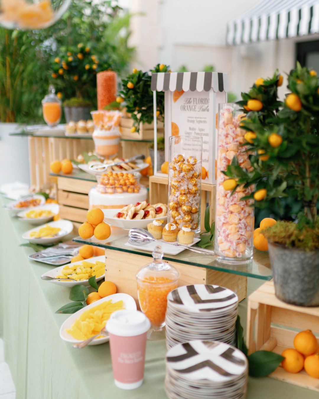 A dessert table decorated with orange-themed treats, plates, cups, glass jars of candy, and potted orange trees, arranged on wooden crates and a green tablecloth.