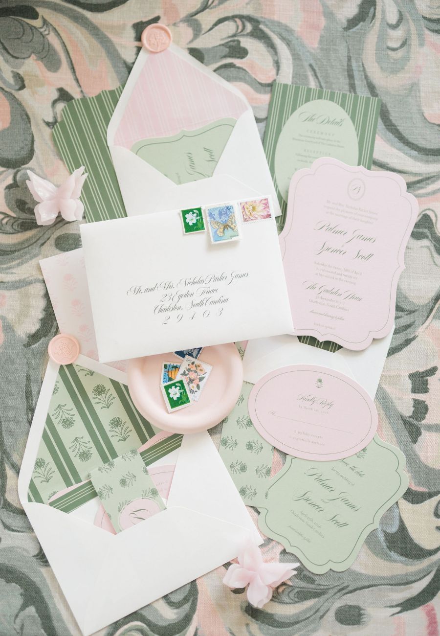 A flat lay of pastel wedding invitations, envelopes with stamps, green and pink cards, and decorative items on a marbled background.