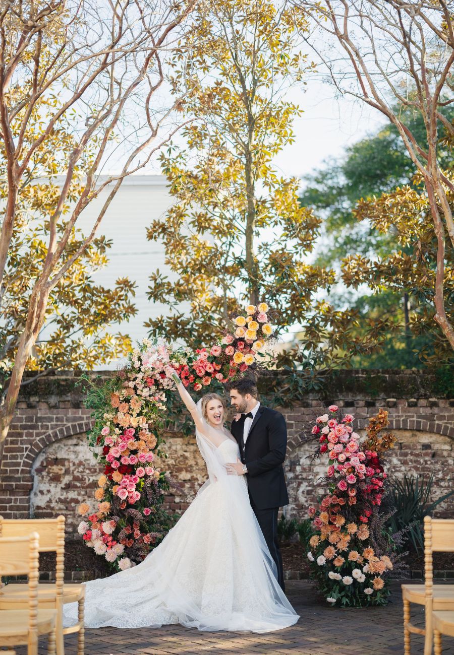 Bride and groom stand together under a floral arch outdoors, surrounded by trees and empty wooden chairs, on their wedding day.