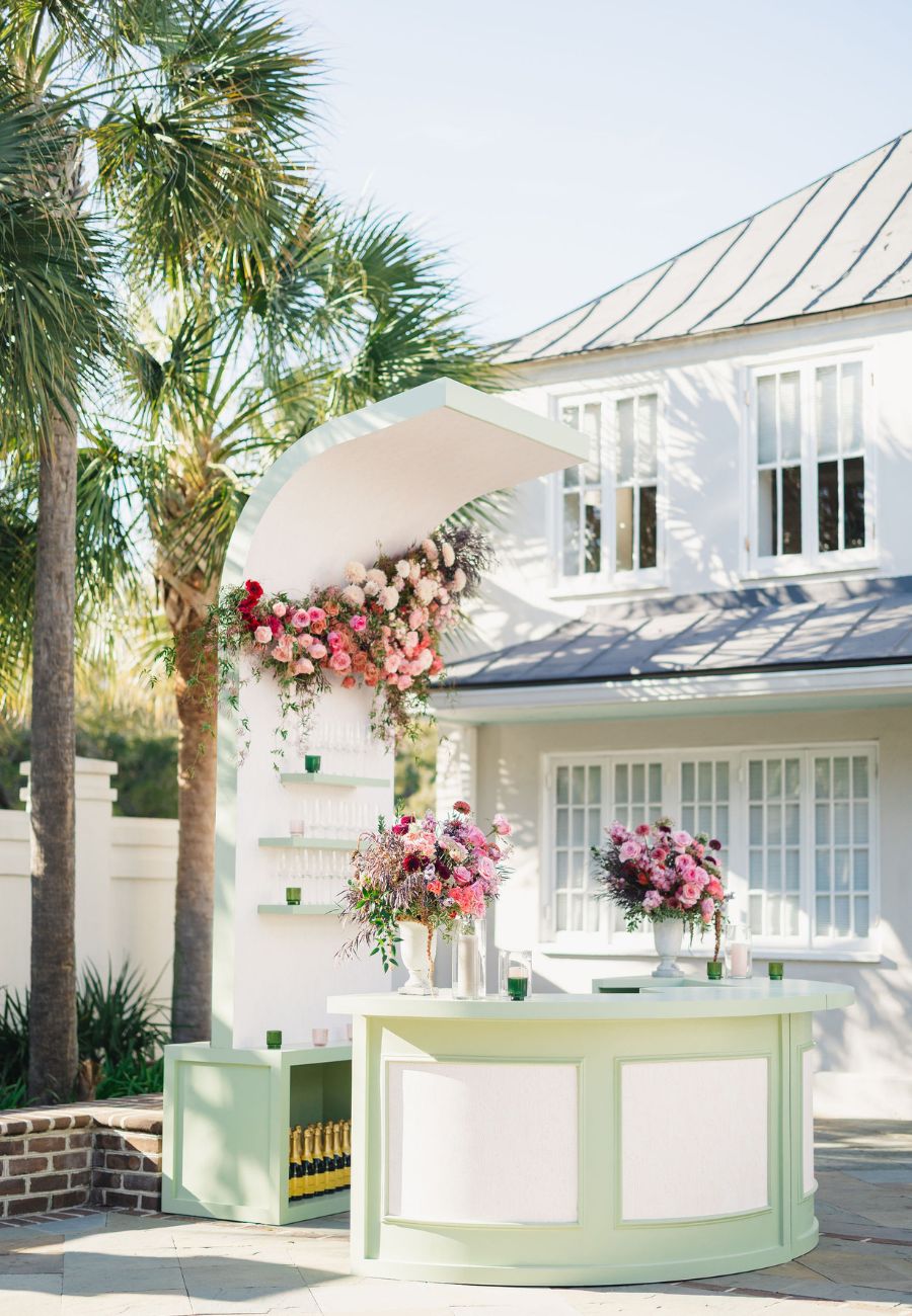 Outdoor bar setup with pastel green and white counter, floral arrangements, and decorative arch, positioned in front of a white house with palm trees in the background.