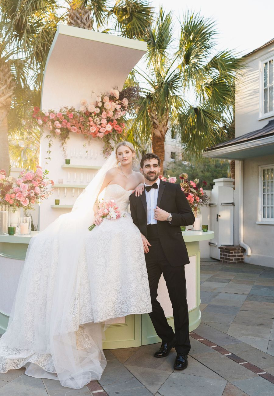 A bride in a white gown and a groom in a black tuxedo pose together outdoors beside a floral-decorated white structure with palm trees in the background.