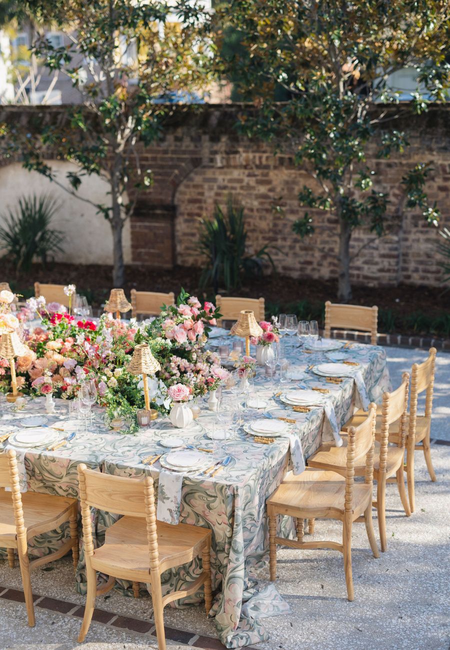 A long outdoor table set with floral arrangements, plates, glasses, and wooden chairs, placed in a garden with trees and a brick wall in the background.