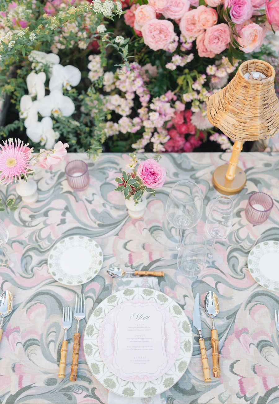 A table set for a meal with floral-patterned plates, cutlery, a small wicker lamp, pink candles, and pink flowers, against a backdrop of various blooming flowers.