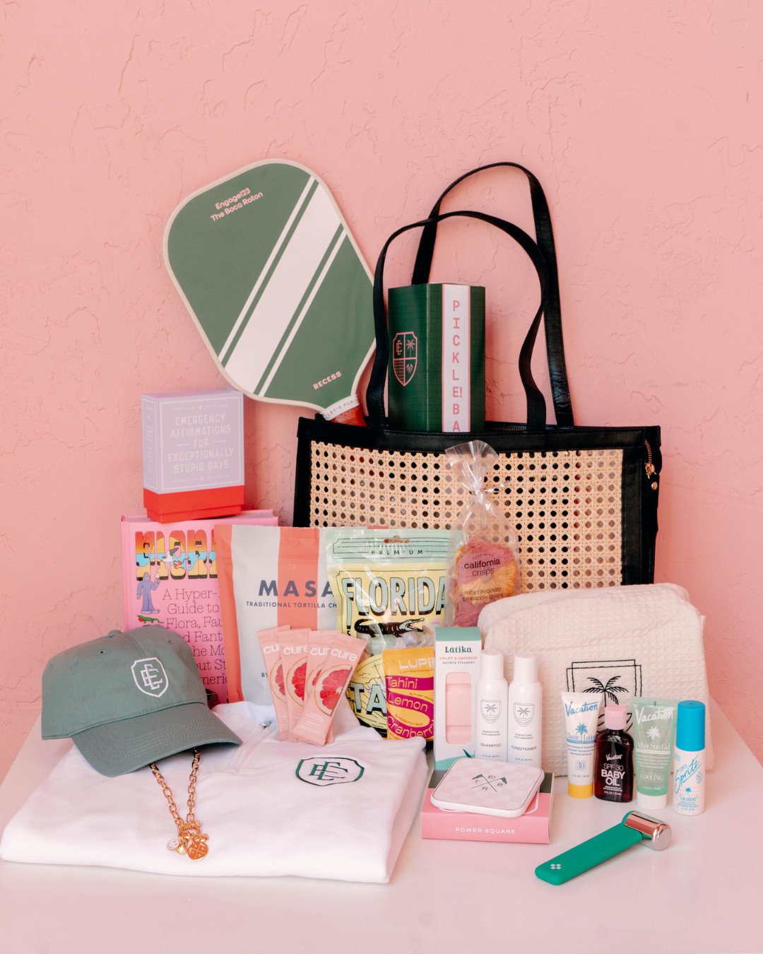 A collection of assorted gift items including a tote bag, paddle, cap, skincare products, snacks, books, and accessories arranged against a pink background.