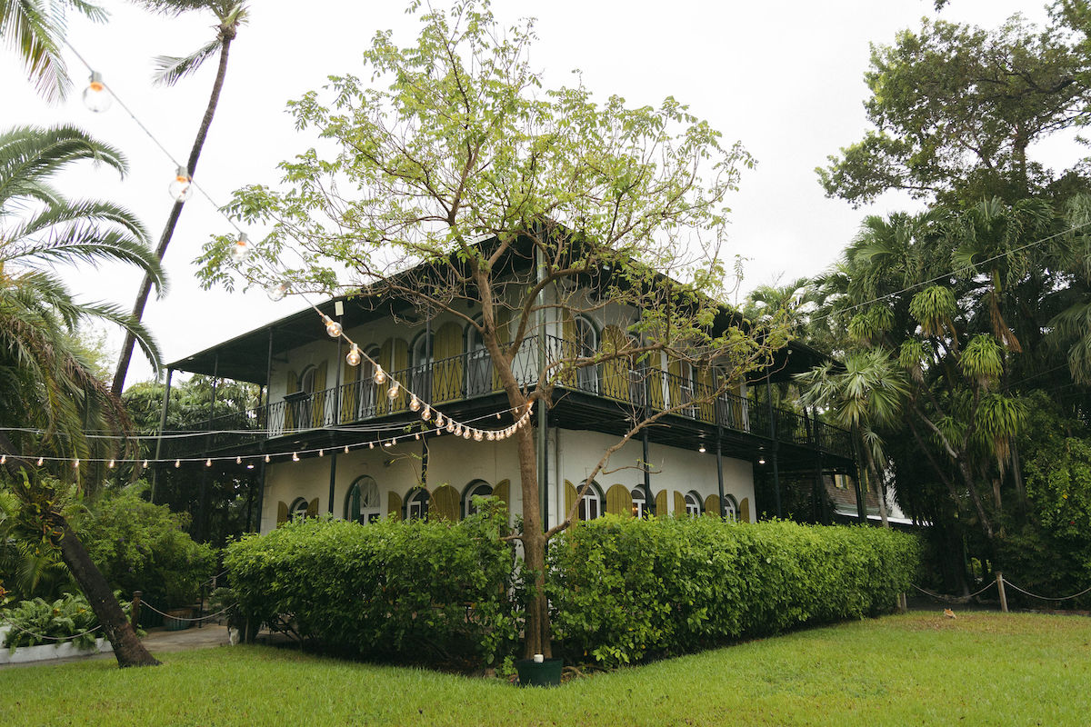 A two-story house with yellow-trimmed windows and a wraparound balcony is surrounded by lush greenery and palm trees.