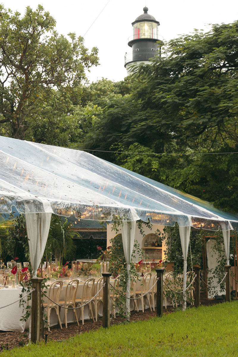A clear tent set up for an outdoor event with tables and chairs, surrounded by greenery, and a lighthouse visible in the background.