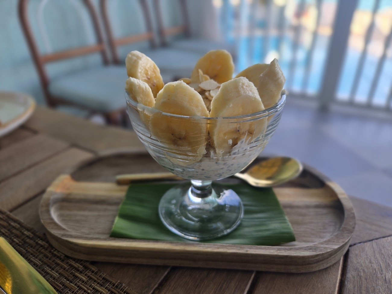 A glass bowl filled with sliced bananas sits on a wooden tray with a green leaf and a gold spoon, on a table near chairs and a railing.