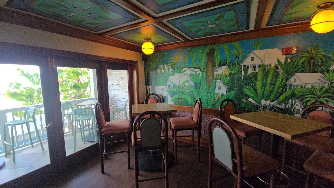 Small dining area with several tables and chairs, colorful mural of tropical scenery on the wall, and glass doors leading to an outdoor balcony.