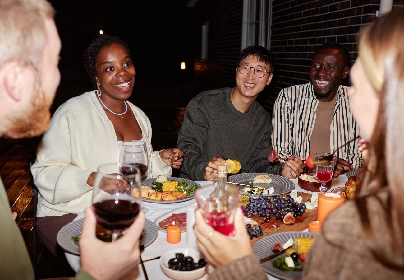 Five people sit around a table outdoors, sharing food and drinks, smiling and engaging in conversation during an evening meal.