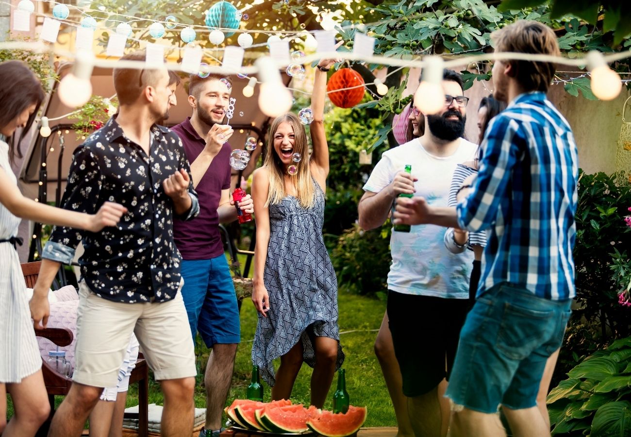 A group of young adults enjoying an outdoor party, laughing and dancing, with string lights overhead and sliced watermelon on the table.