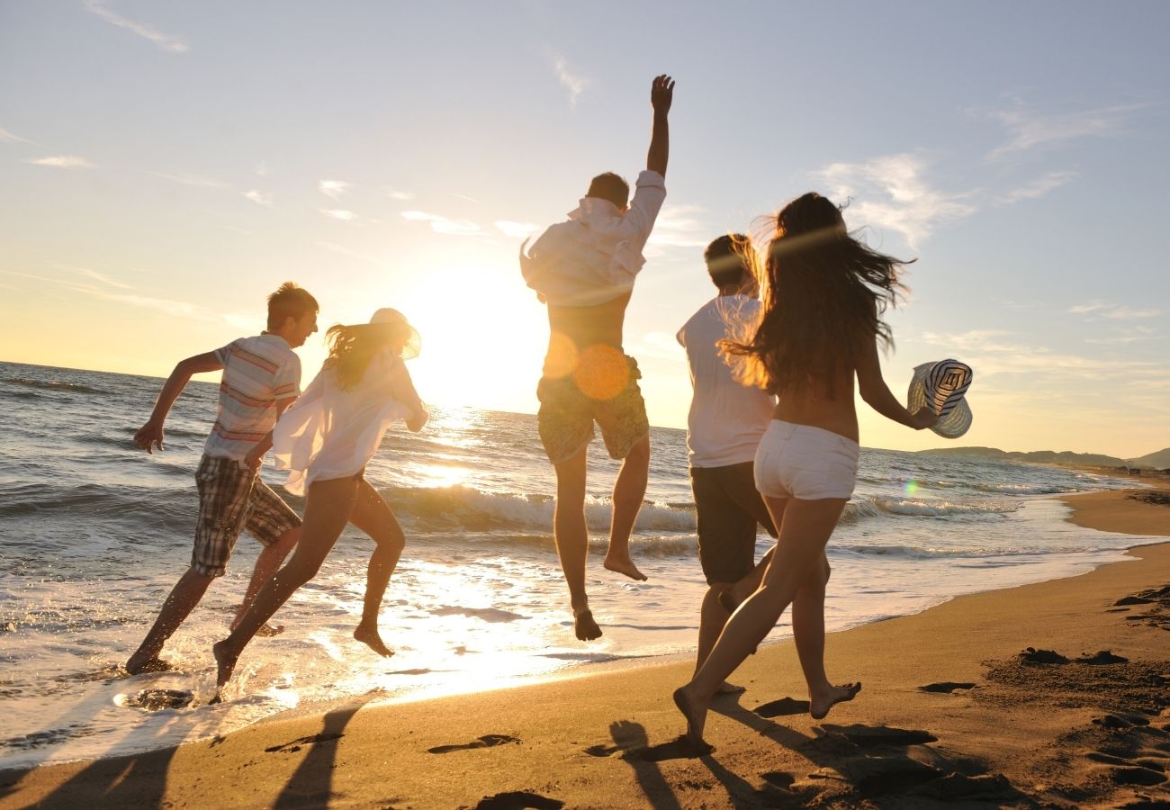 Five people run and jump along the beach at sunset, with waves in the background and sunlight shining behind them.