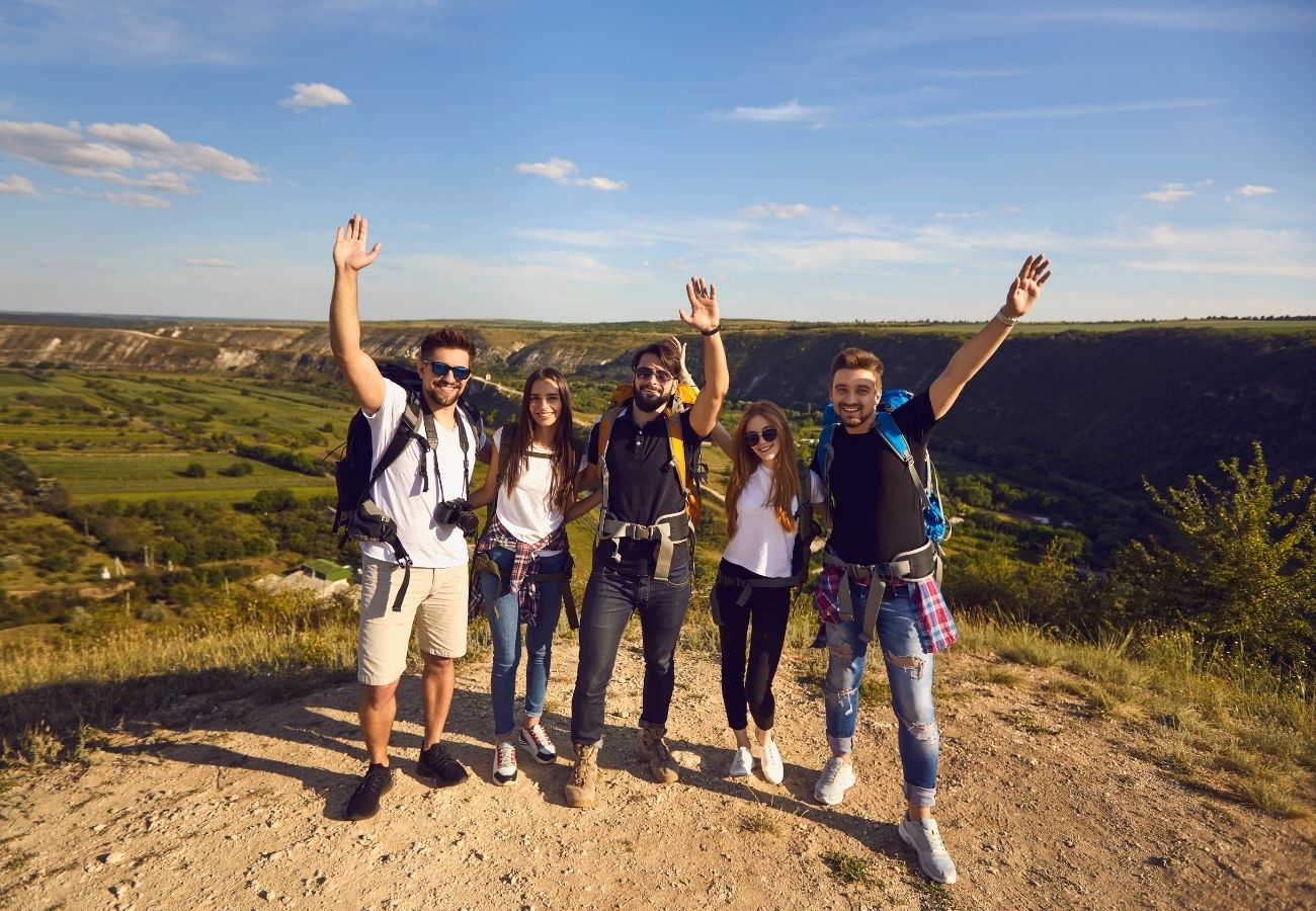 Five people with backpacks stand on a hilltop, smiling and raising their arms, with a scenic valley and blue sky in the background.