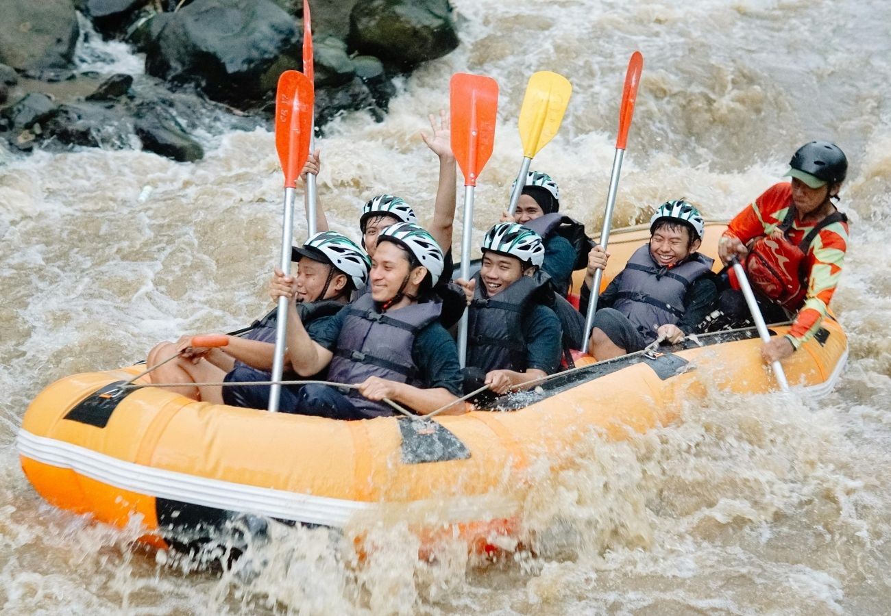 A group of people wearing helmets and life jackets paddle an inflatable raft through rough, muddy river water.