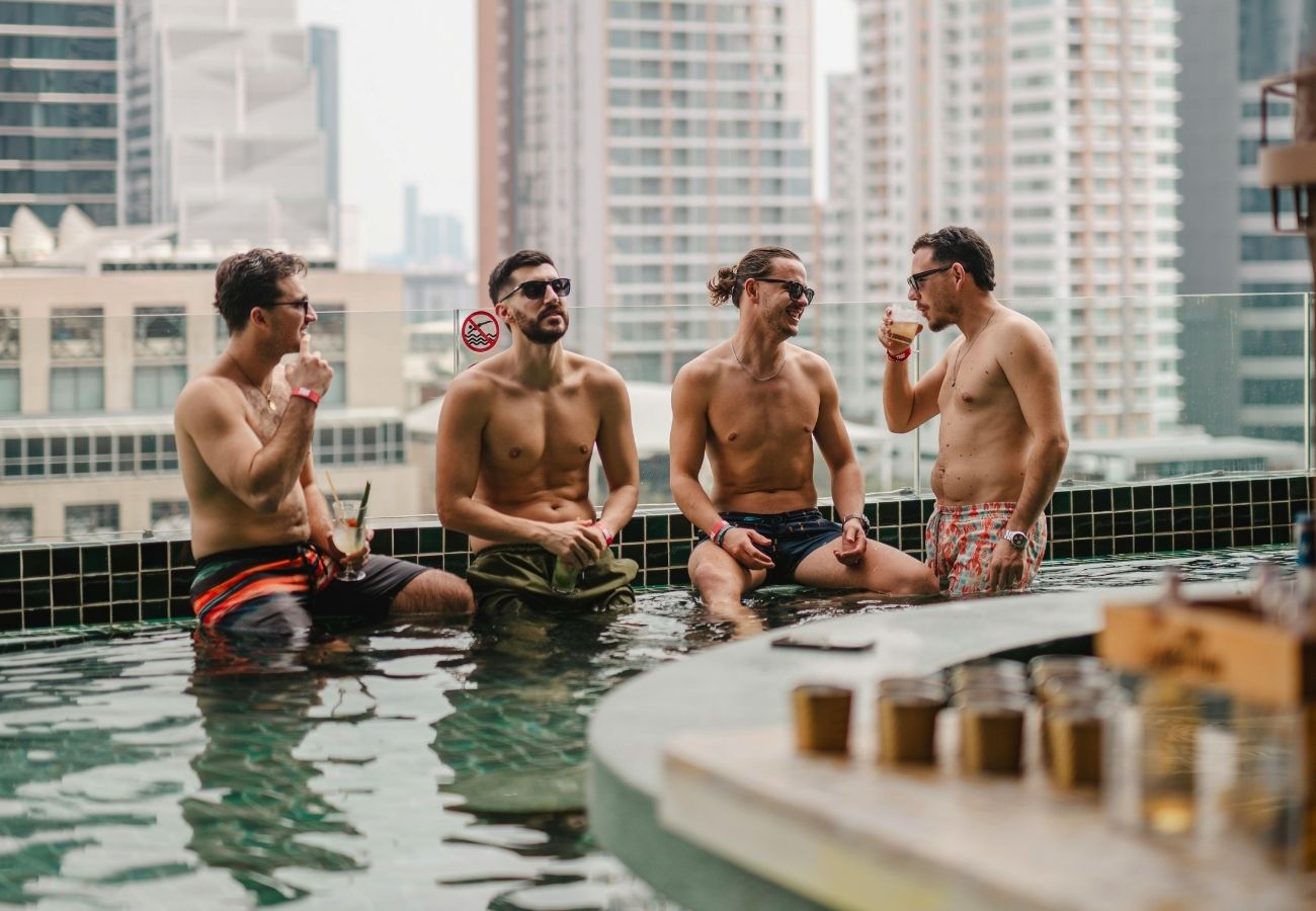 Four men in swim shorts sit and stand in a rooftop pool, drinking and talking, with tall city buildings in the background.