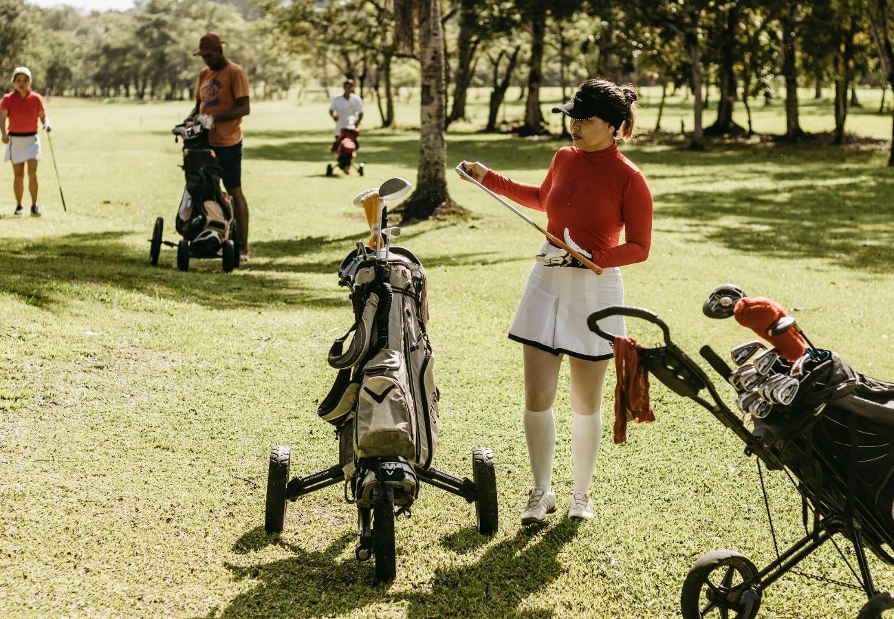 A woman in a red top and white skirt stands next to a golf bag on a golf course, reading a scorecard, with other golfers in the background.