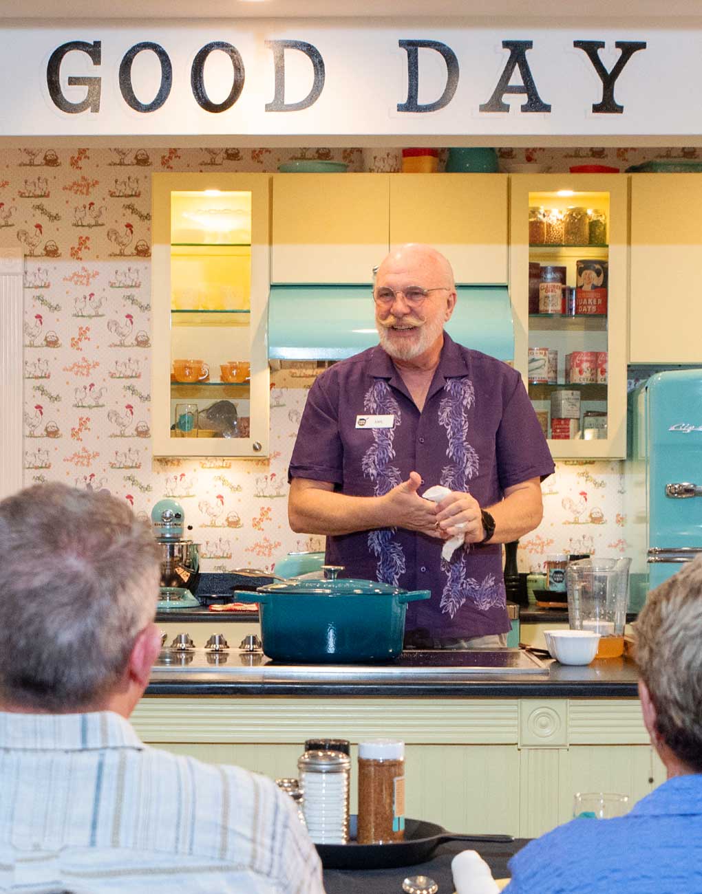 A man stands behind a kitchen counter giving a cooking demonstration to an audience; a sign above reads "GOOD DAY."