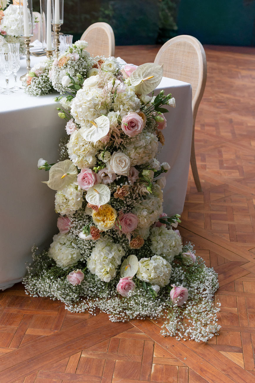 A floral arrangement of white hydrangeas, roses, and baby's breath cascades from a table to the floor beside two chairs on a parquet wood floor.