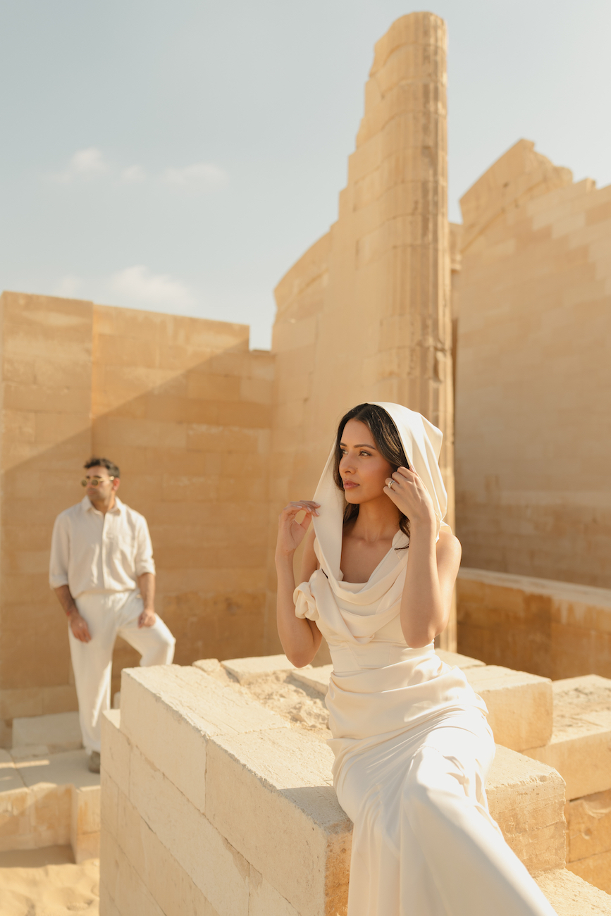 A woman in a white outfit sits on a stone structure, adjusting her hood, while a man in white stands in the background among beige ruins under a clear sky.