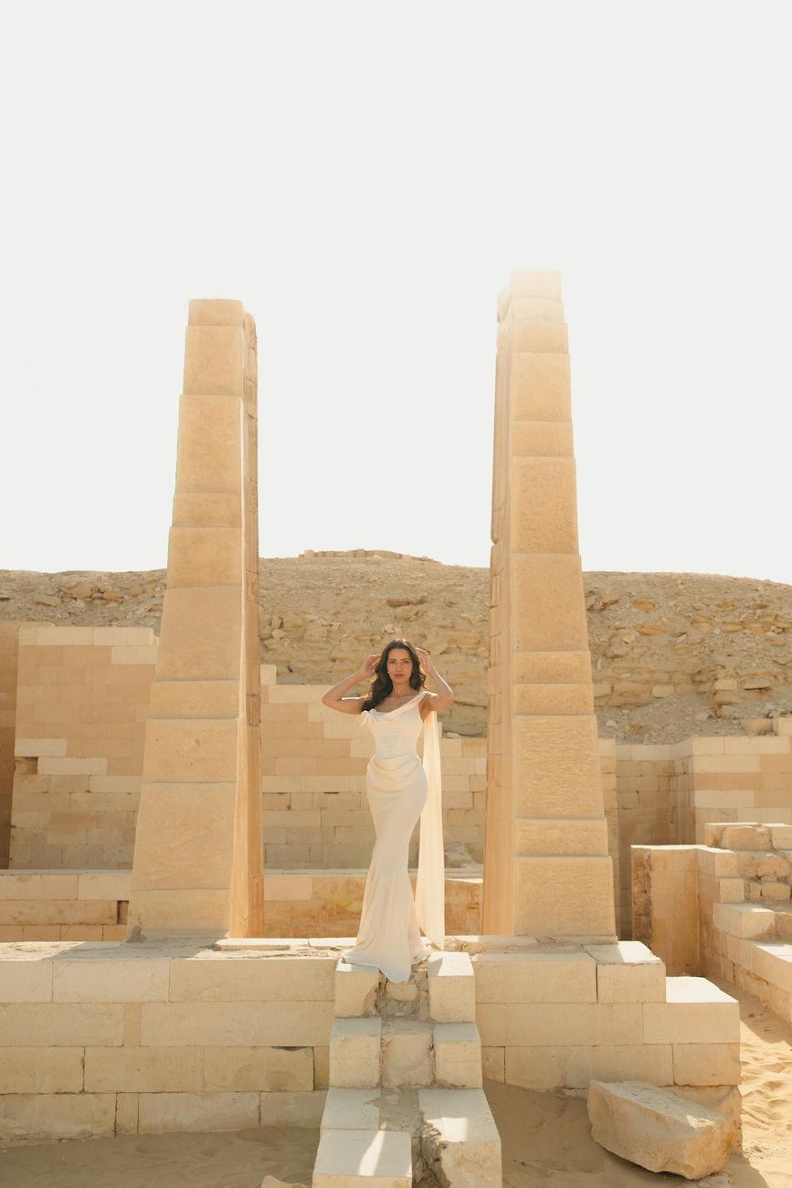 A woman in a white dress stands between two tall ancient stone pillars at an archaeological site in a desert landscape.