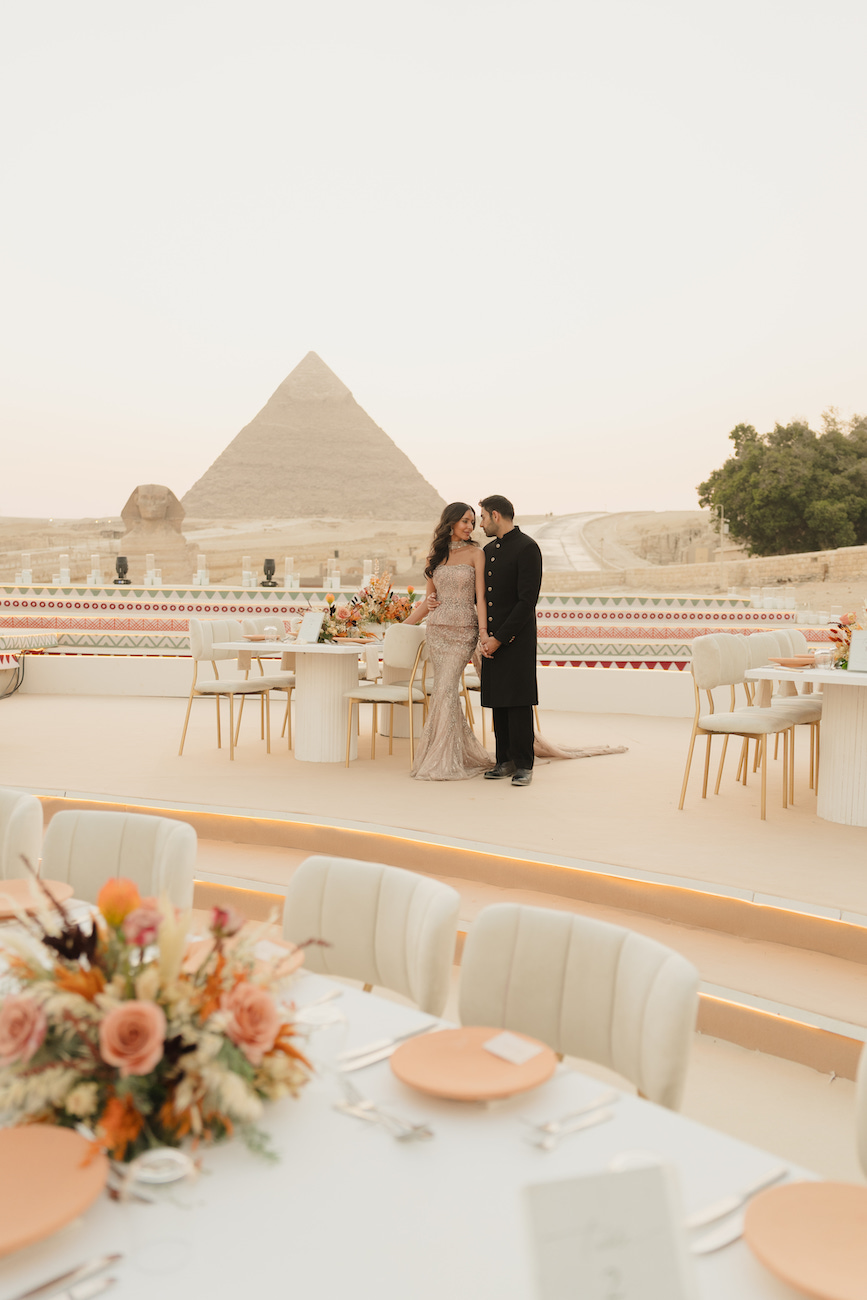 A couple stands close together at an elegant outdoor event with decorated tables and the Pyramid of Giza visible in the background.