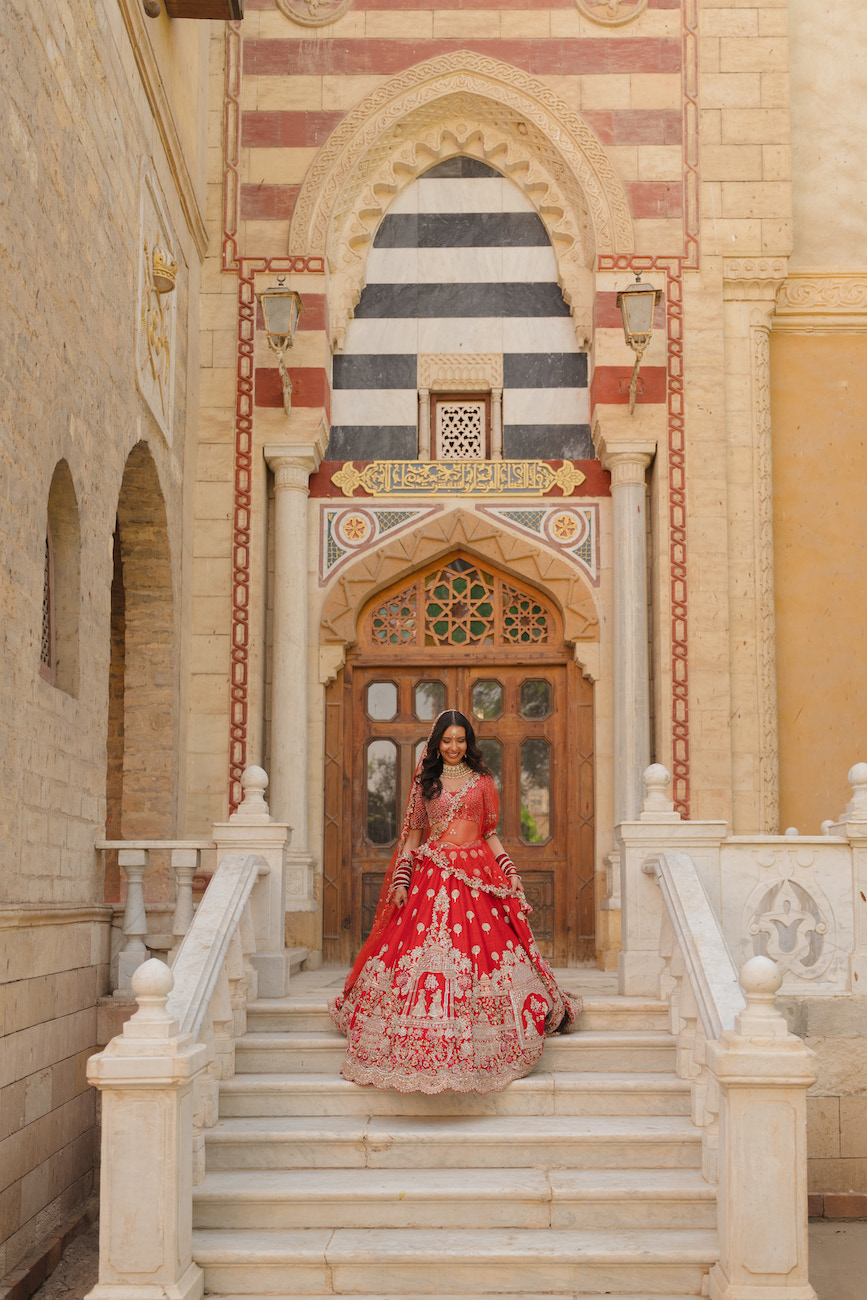 A woman in a red and gold traditional outfit stands on white stone steps in front of an ornate arched doorway with geometric designs.