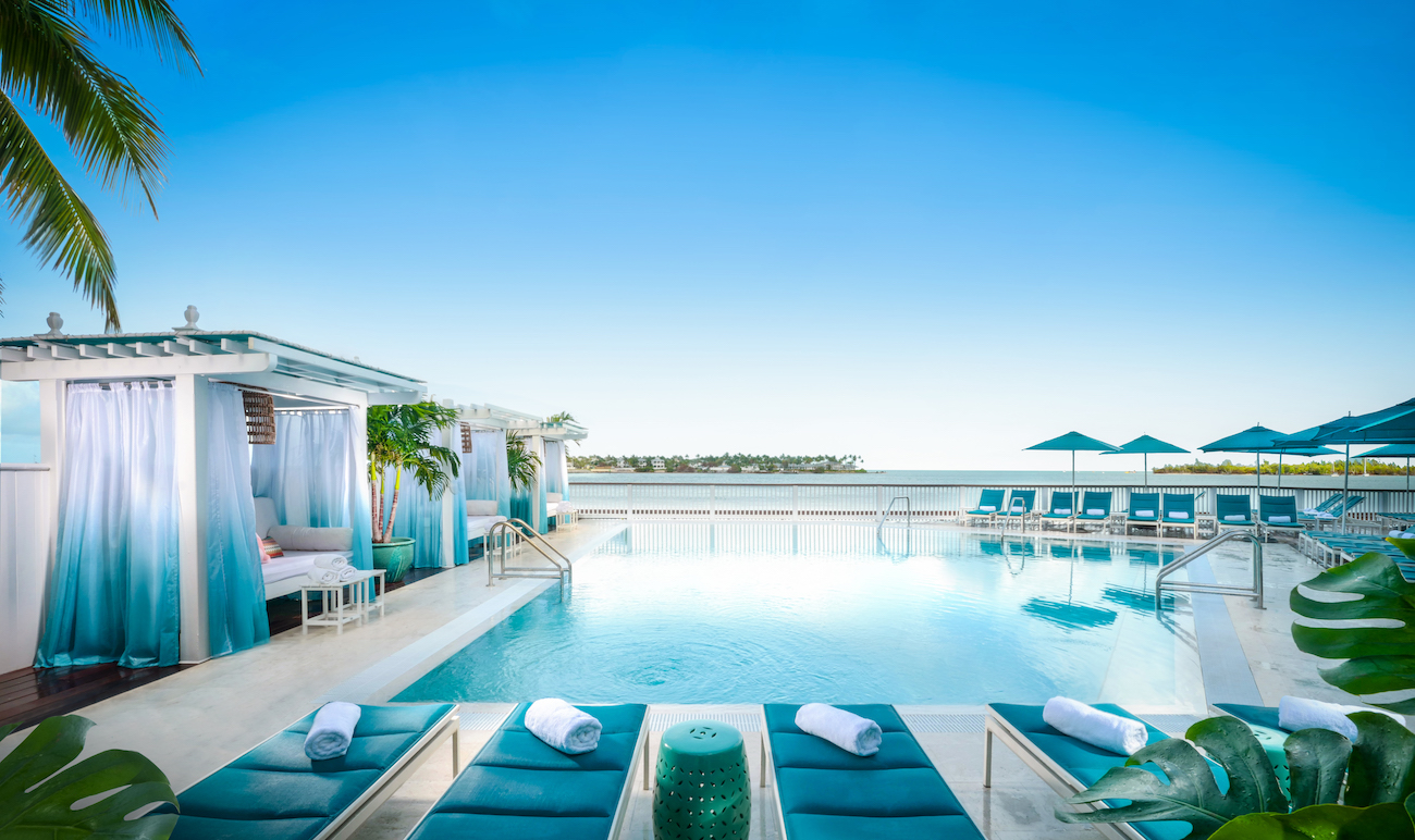 A rooftop pool area with lounge chairs, cabanas, and umbrellas overlooks the ocean under a clear blue sky.