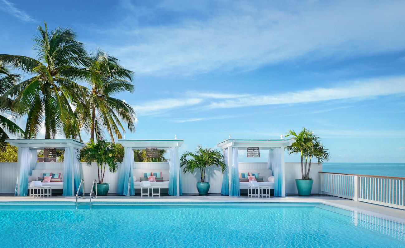 Three white cabanas with lounge chairs are lined up beside a swimming pool, with palm trees and a clear blue sky in the background.