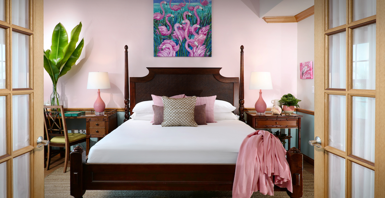 A neatly made bed with pink accents is centered in a bedroom with light pink walls, wooden furniture, and a flamingo painting above the headboard.