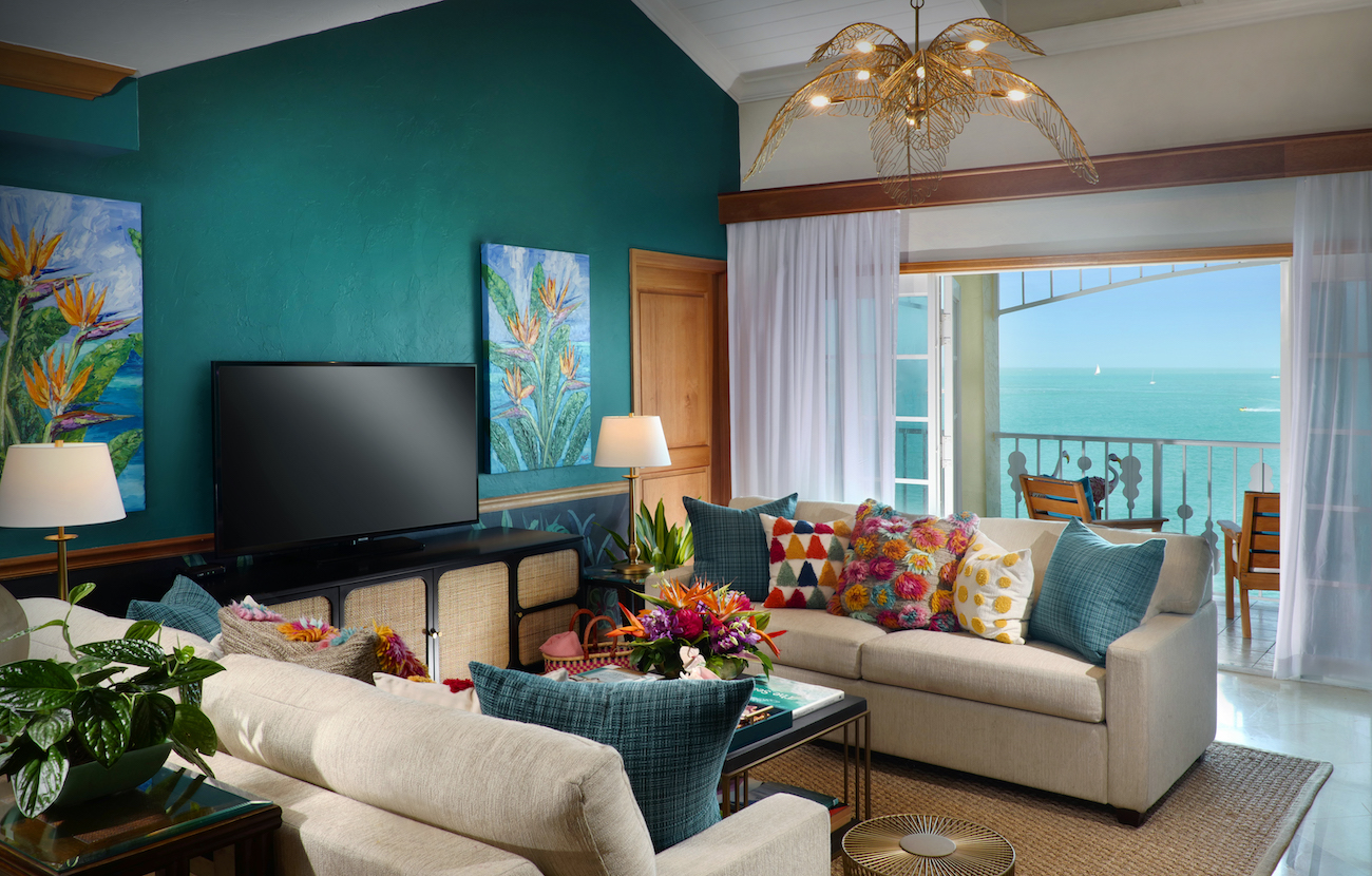 Bright living room with teal accent wall, colorful pillows, two sofas, a TV, and large window opening to a balcony with an ocean view.