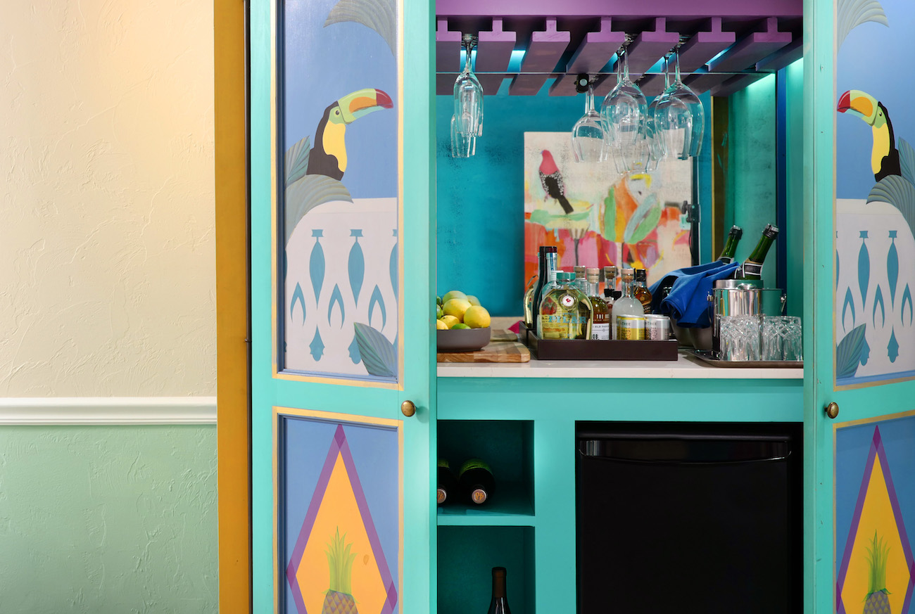A colorful cabinet with painted toucans holds a small bar setup, including bottles, a tray, lemons, glasses, and a mini fridge.