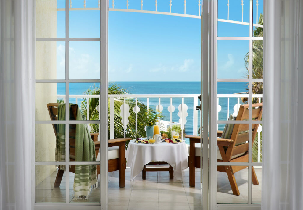 Two wooden chairs and a small table with breakfast items overlook the ocean from a balcony, viewed through open glass doors on a sunny day.