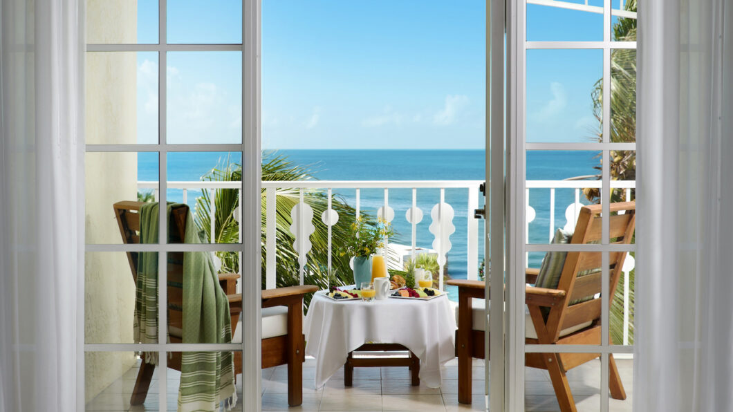 Two wooden chairs and a small table with breakfast items overlook the ocean from a balcony, viewed through open glass doors on a sunny day.