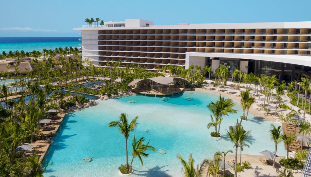 A modern beachfront resort with a large lagoon-style pool, palm trees, and a multi-story hotel building under a clear blue sky.