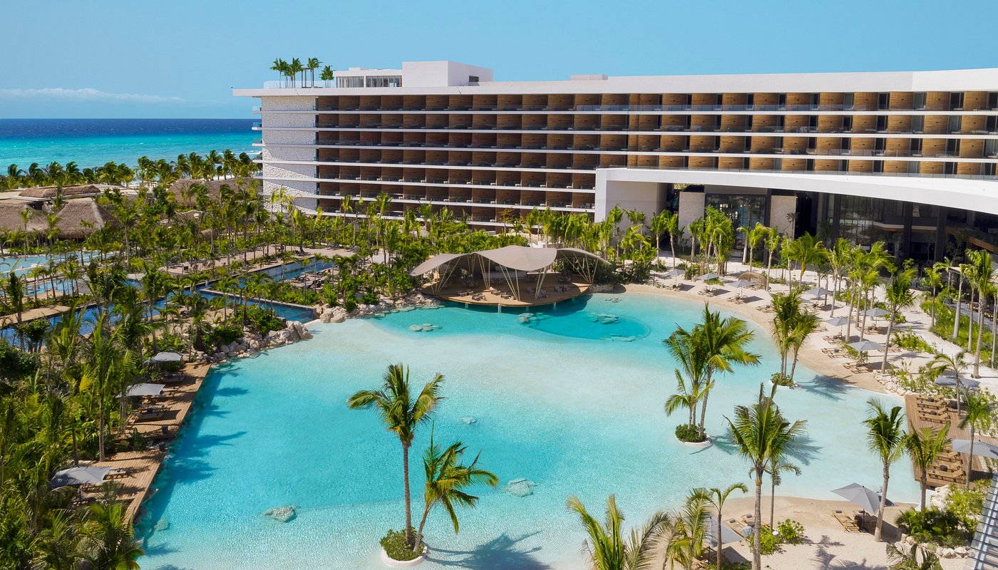 A modern beachfront resort with a large lagoon-style pool, palm trees, and a multi-story hotel building under a clear blue sky.