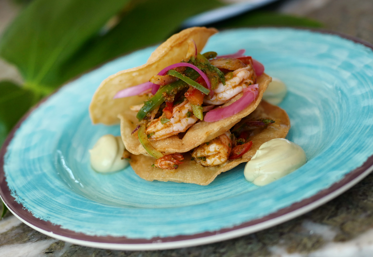 A plated dish with stacked tostadas, topped with shrimp, sliced red onions, and peppers, garnished with dollops of green sauce on a blue and white plate.