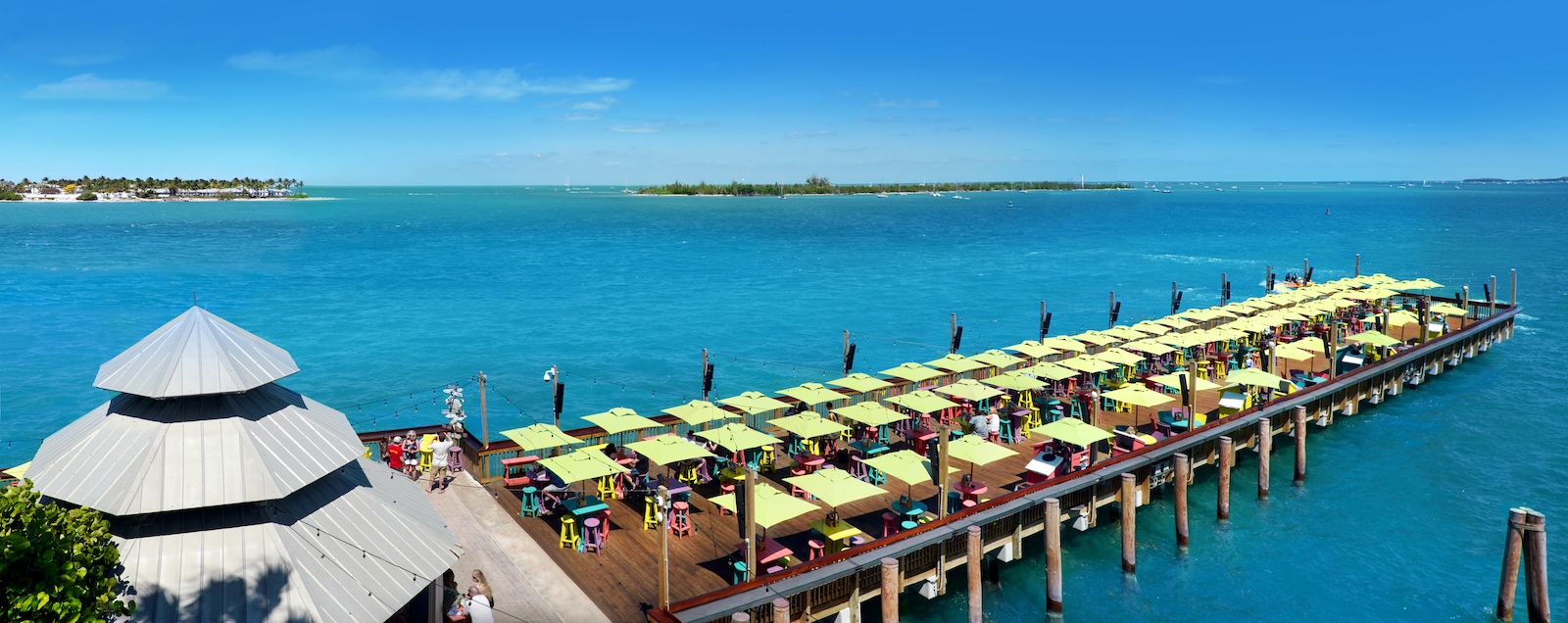 A long pier with rows of yellow-green umbrellas over tables stretches into turquoise water, with a building on the left and islands visible in the distance under a clear blue sky.
