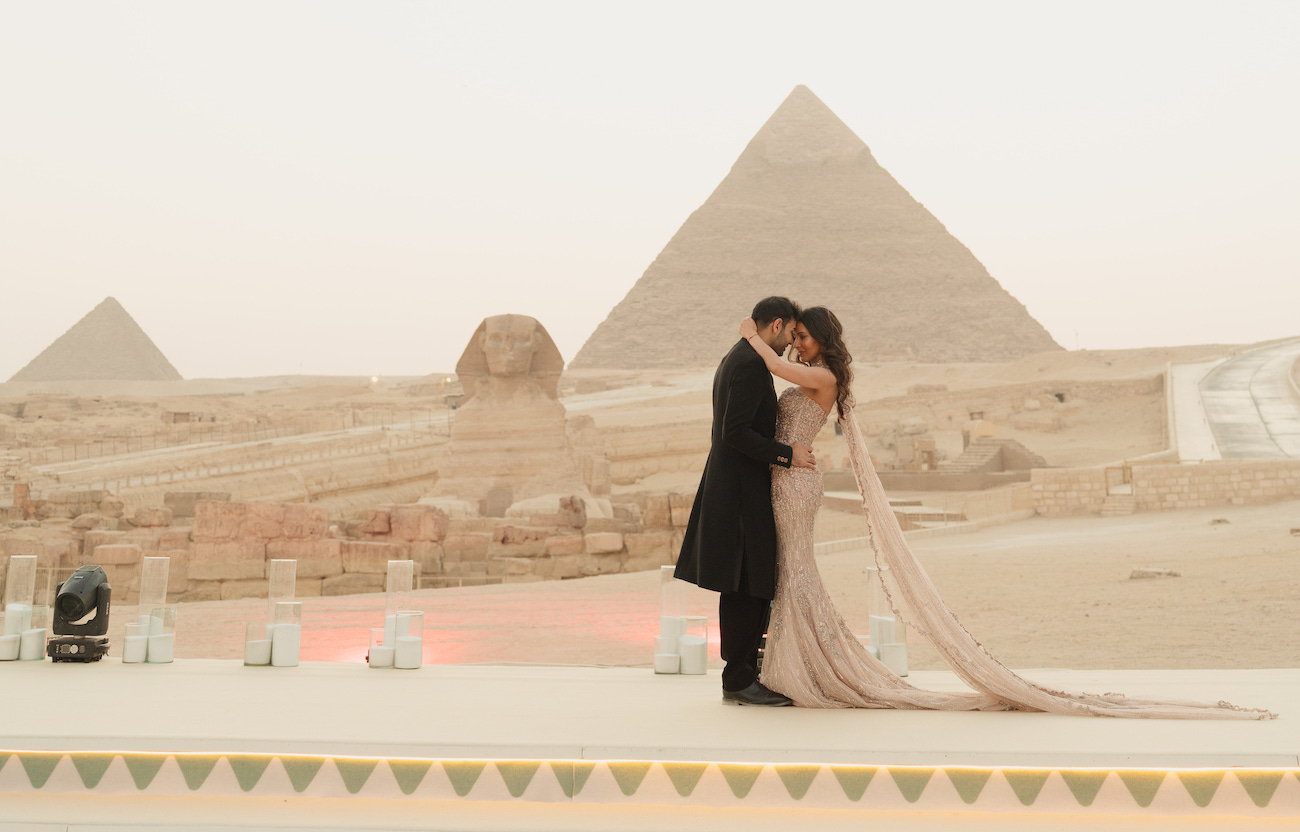 A couple dressed formally embraces on a platform near the Sphinx and a pyramid in Egypt, with soft lighting and candles arranged along the edge.