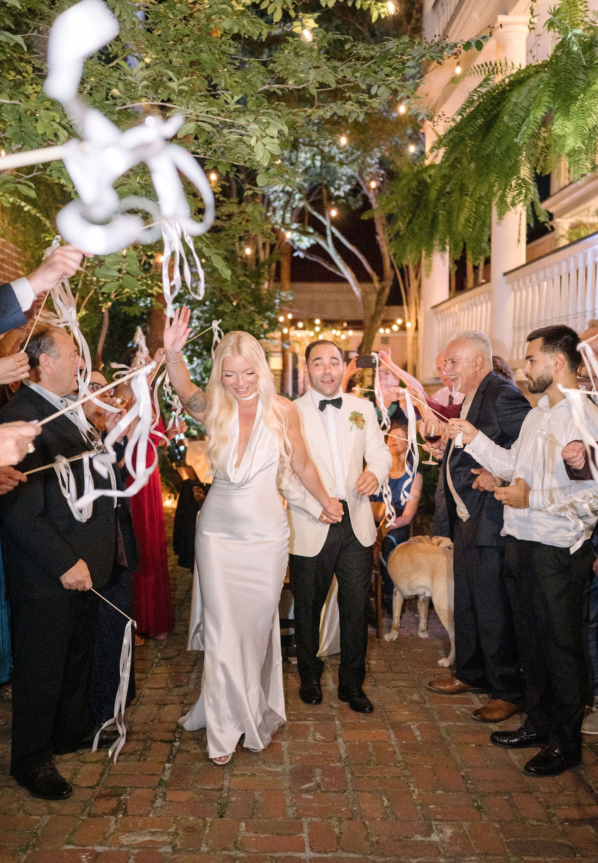 A bride and groom walk down a brick pathway at night, surrounded by guests waving streamers; string lights and greenery decorate the scene.