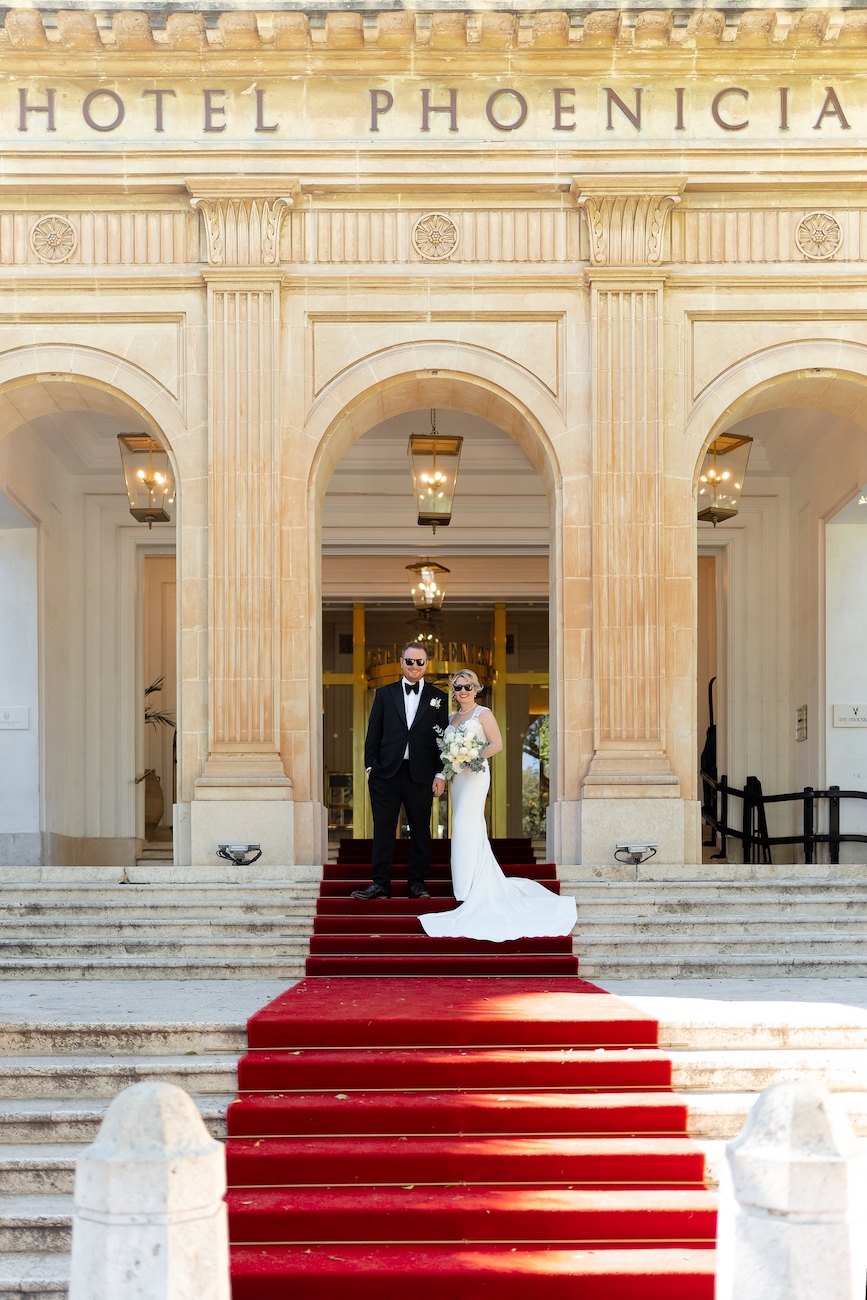 A bride and groom stand on a red carpeted staircase in front of the entrance to Hotel Phoenicia.
