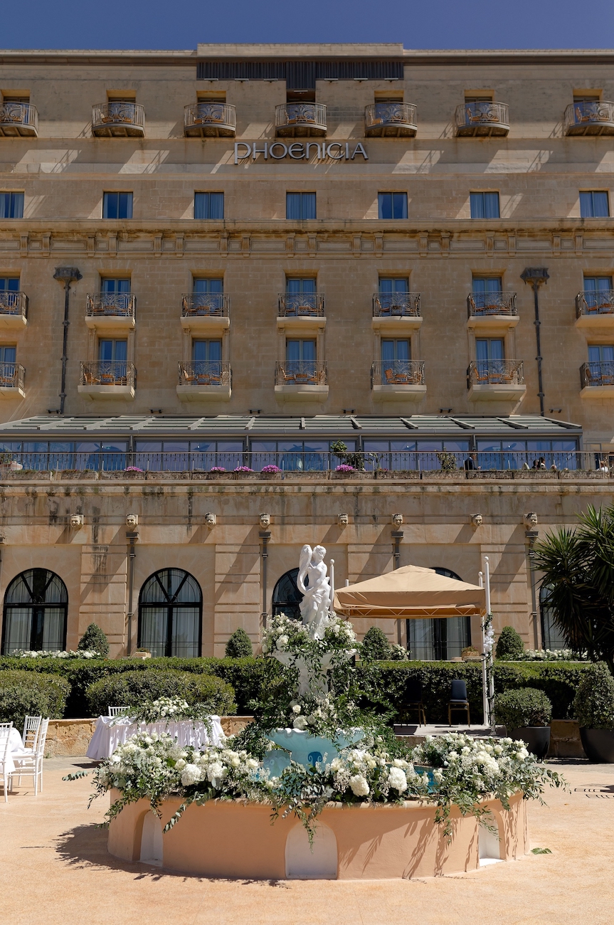 A stone fountain with a white statue and flower arrangements is in front of a large, multi-story hotel building with many windows and balconies.
